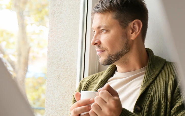 Eine männliche Person mit Tasse in der Hand schaut an die Wand gelehnt aus einem Fenster