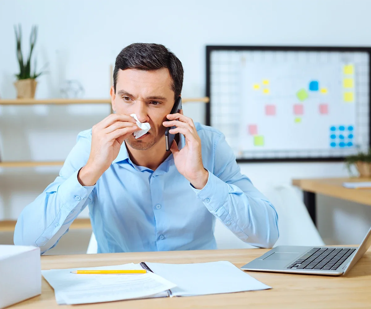 Eine männliche Person an einem Schreibtisch sitzend mit Telefon in der einen und in der anderen Hand ein Taschentuch an der Nase