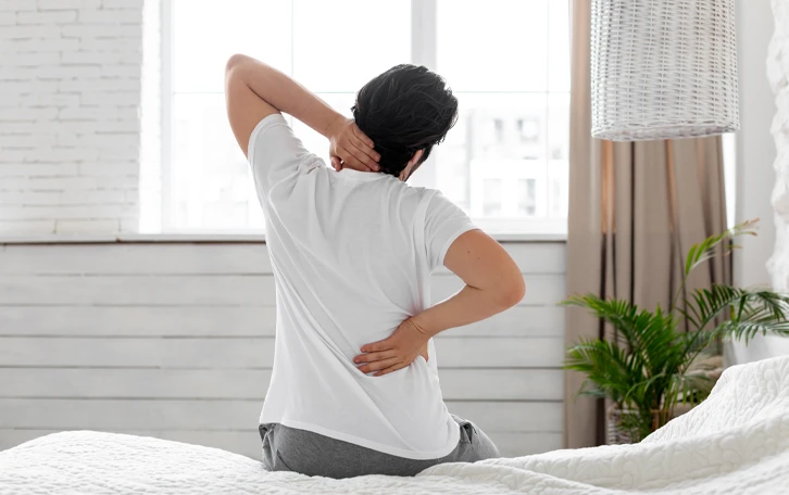 Eine männliche Person auf einer Bettkante sitzend hält sich den Nacken- und Lendenbereich