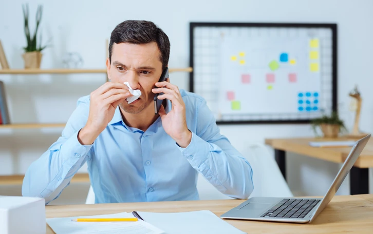 Eine männliche Person an einem Schreibtisch sitzend mit Telefon in der einen und in der anderen Hand ein Taschentuch an der Nase