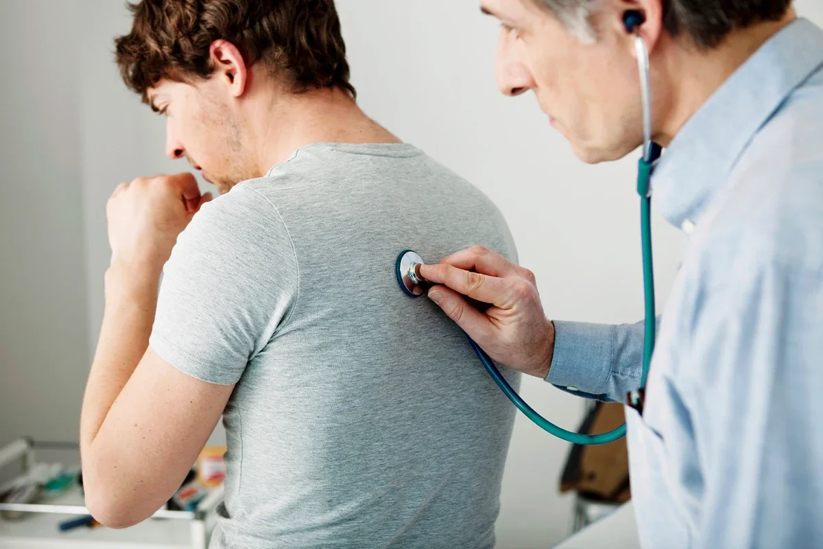 Eine männliche Person wird von einem Arzt mit einem Stetoskop auf dem Rücken abgehört